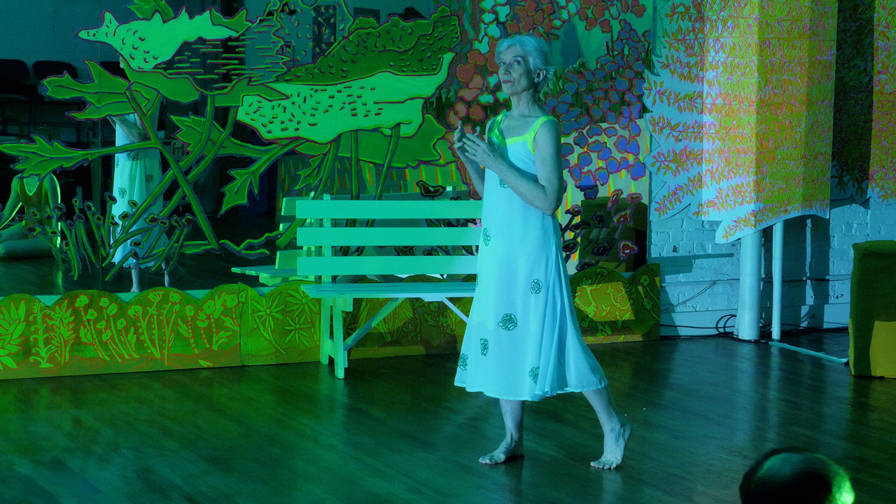Gray haired woman dressed in a long flowing light colored dress standing in front of a white garden bench that's situated next to the brightly colored painted mirror.